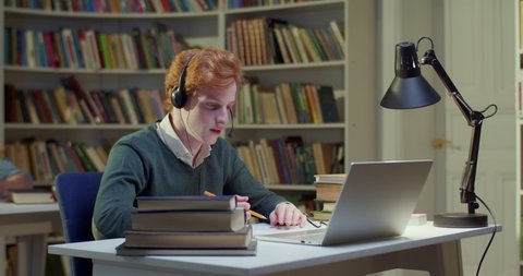 Caucasian red-haired male in headset talking, writing and videochatting on laptop in library. Young man speaking, taking notes and having videochat online via webcam. Video call. Noting information.