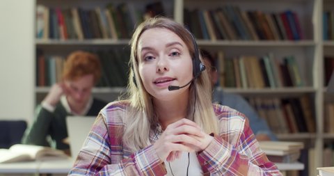 Pretty blonde Caucasian female in headset talking, videochatting for education vlog in library. Woman speaking, having videochat. Video call in bibliotheca. Tutoring. Educational vlogging. Close up.