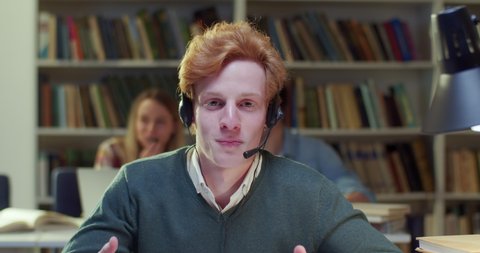 Red haired Caucasian male in headset talking, videochatting for education vlog in library. Young man speaking, having videochat. Video call in bibliotheca. Tutoring. Educational vlogging. Close up.