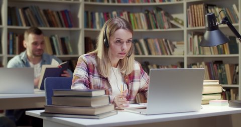 Pretty blond female in headset talking, writing and video chatting on laptop in library. Attractive woman speaking, taking notes and having video chat online via webcam. Video call. Noting information.