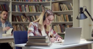 Beautiful blonde female in headset talking, writing and videochatting on laptop in library. Woman speaking, taking notes and having videochat online via webcam. Video call. Noting information.