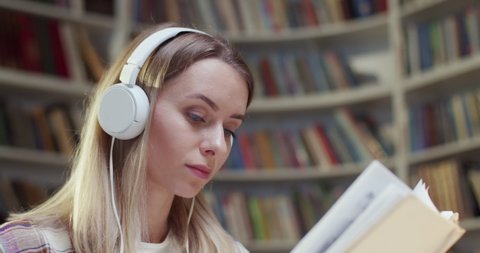 Close up of beautiful young Caucasian woman in headphones reading book and listening to music in library. Educational concept in bibliotheca. Female student listen to song and studying.
