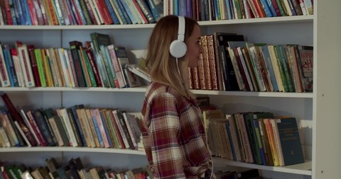 Pretty young Caucasian female student in headphones choosing textbook in library and sitting down at table to study. Blonde woman taking out book from shelf in bibliotheca and reading.