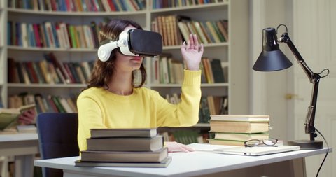 Caucasian female student in VR headset sitting in library space, scrolling and moving hands in air and having virtual reality experience. Metaverse studying. Study in virtuality concept.