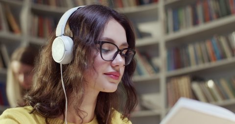 Close up of beautiful young Caucasian woman in glasses and headphones reading book and listening to music in library. Educational concept in bibliotheca. Female student listen to song and studying.