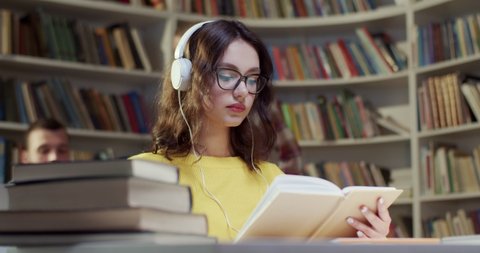 Beautiful young Caucasian woman in glasses and headphones reading book and listening to music in library. Educational concept in bibliotheca. Female student listen to song and studying.