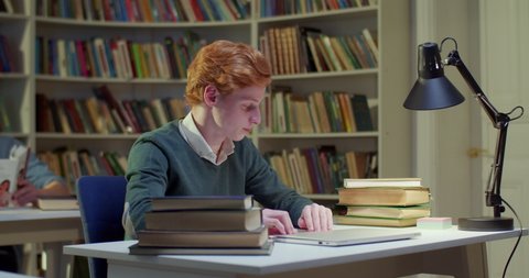 Young Caucasian male student choosing book at table in library and opening to read. Red-haired man taking textbook and reading it. Study concept in bibliotheca. Preparing for exams.
