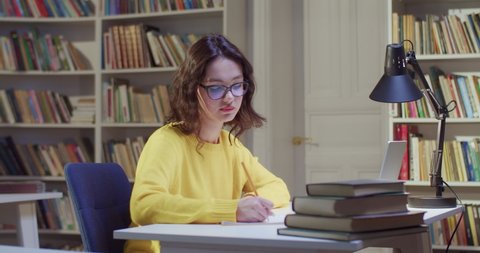 Young Caucasian beautiful female student studying in library, resting and stretching her neck. Break in study and rest in bibliotheca. Woman in glasses learning and working with books.