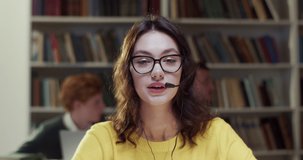 Young Caucasian female in headset and glasses talking and videochatting for education vlog in library. Woman speaking and having videochat. Video call in bibliotheca. Tutoring. Educational vlogging.