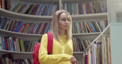 Portrait of young beautiful Caucasian female student in glasses and with red backpack standing in library, choosing textbook to read and then smiling to camera. Taking out book from shelf.