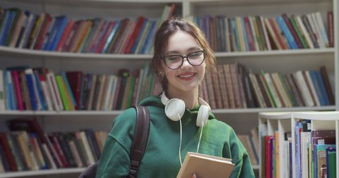 Portrait shot of young Caucasian pretty female student in glasses smiling to camera and holding textbook in library. Woman with book in hands standing at bookshelves. Literature concept.