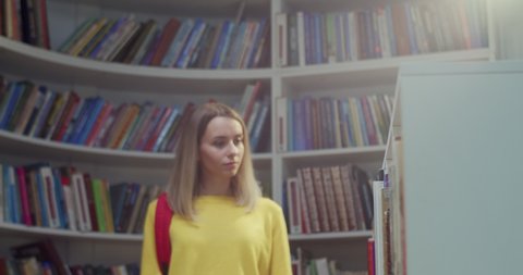 Blonde young beautiful Caucasian female student and with red backpack walking in library, choosing textbook to read and flipping pages. Taking out book from shelf. Literature concept.