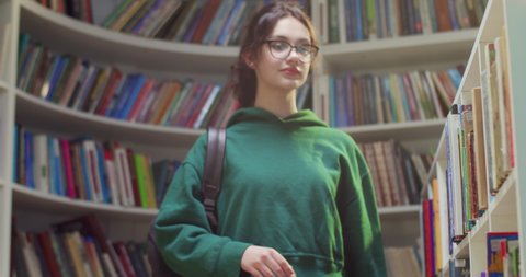 Young beautiful Caucasian female student in glasses and with backpack standing in library, choosing textbook to read. Taking out book from shelf. Literature concept. Close up.