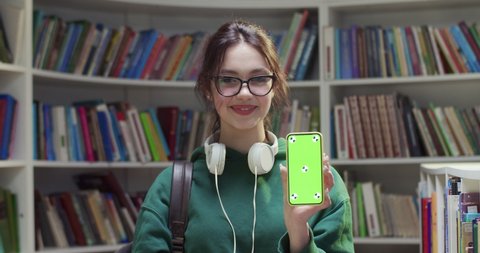 Portrait of young Caucasian pretty female student in glasses smiling and showing smartphone with green screen and tracking motion in library. Chroma key. Woman with mobile phone demonstrating.