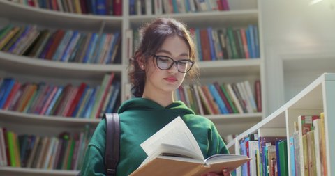 Young beautiful Caucasian female student in glasses and with backpack standing in library, reading textbook and closing it. Putting book on shelf and leaving. Literature concept. Close up.