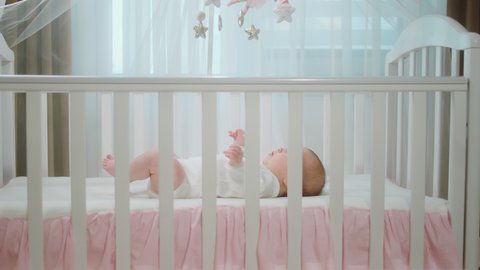 Pretty baby girl very handsome laying down on her baby cot alone playing with hands and legs excited she is concentrated at cot toys looking up