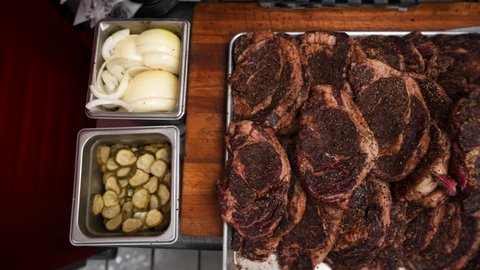 Overhead view of pile of seasoned ribeye steaks on butcher block, cutting board station in steakhouse with pickles, onions, meat cleaver, slider 4K
