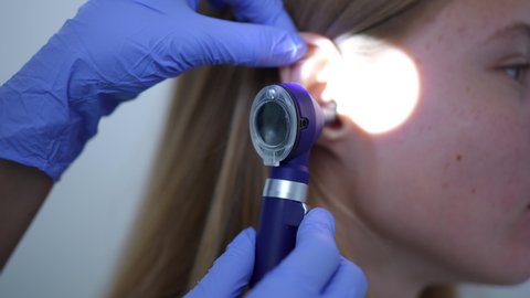 Close-up side view ear of female teenage patient with rack focus to otoscope in doctor hand. Unrecognizable otolaryngologist checking auditory canal and tympanic membrane in hospital indoors