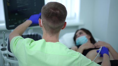 Back view doctor talking pointing at screen doing ultrasound scanning in hospital indoors. Blurred woman in coronavirus face mask lying on examination couch at background. Medicine and gynecology