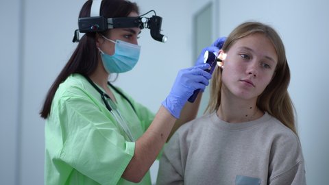 Teenage Caucasian girl sitting in hospital as otolaryngologist with otoscope entering checking ears. Portrait of teen patient and professional female doctor indoors. Medical examination concept
