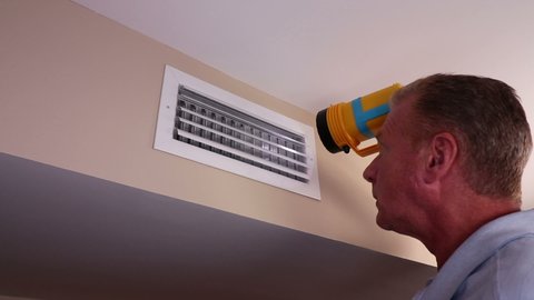 Adult white male with a flashlight inspecting the inside of an air duct in a home wall near a ceiling. Home air vent duct on a wall near a ceiling being inspected by a mature man with a flashlight.