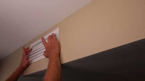 Two male hands closing a rectangle air vent adjustable register blades on a home wall near a ceiling one at a time. Mature male hands shutting the air duct register blades one at a time for air flow