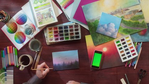 Artist painting landscape with watercolors and brush top view. Art and inspiration, designer. Workspace of creative person using phone with chroma key, talented human. Leisure and hobby concept.