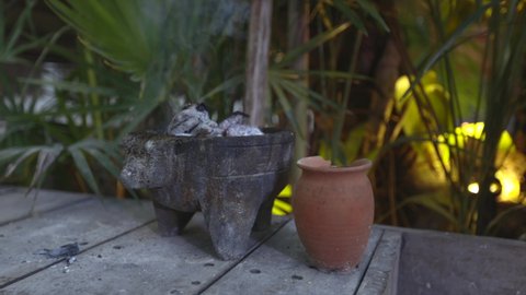 Traditional Incense burner with burning coal and strong smoke on wood floor in a tropical garden with organ warm light and green plants around