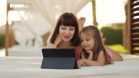 Happy smiling mom holding tablet pc with her little kid girl, while resting in the outdoor gazebo near swimming pool having a video call waving their hands. Vacation gadgets and technology concept