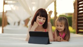 Relaxed young woman and her daughter watching movie or having video chat on digital tablet while lying in the resort gazebo near pool. Happy mother and her daughter sunbathing and holding tablet pc