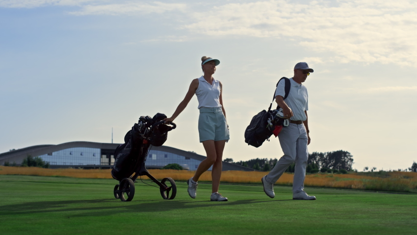 Two golfers walk fairway at sunset. Rich couple carry sport equipment clubs in trolley bag. Smiling successful partners talking discussing golf game on field course landscape. Luxury weekend concept. Royalty-Free Stock Footage #1089897369