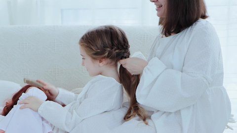 Young happy mother and little cheerful daughter sits on the couch while mom braids child's hair, side view 4k video