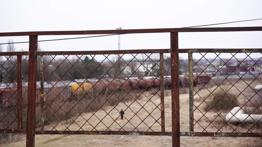 Rusty metal fence overlooking an abandoned railway yard with old freight train wagons in the distance. A small figure walks across the overgrown field below. The scene shows industrial decay, post-Sov