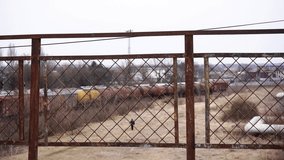 Rusty metal fence overlooking an abandoned railway yard with old freight train wagons in the distance. A small figure walks across the overgrown field below. The scene shows industrial decay, post-Sov - Powered by Shutterstock - Get 15% off with code: PIKWIZARD15