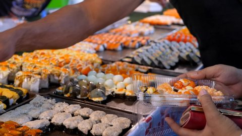 Man Hand Choosing and Imposing Fresh Sushi in Plastic Bag from Night Market Stand. People Buy Japanese Food at Evening. Street Store with Sushi and Sashimi Assortment. Asian Food in Thailand, Phuket