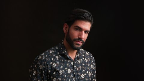 Close up studio portrait of handsome seductive confident intriguing masculine man in floral long-sleeved shirt touching his shiny healthy hair. Black background. High quality 4k footage
