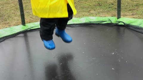 A little girl in blue rubber boots and a yellow raincoat jumps on a trampoline during the rain, splashes, slow motion
