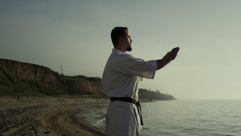 Brutal athlete making meditation exercises on sandy beach summer evening. Karate fighter perfecting combat technique at sunset outdoors. Focused sportsman practicing martial arts at sunlight.