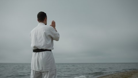 Unknown karate fighter practicing attacks in overcast weather by day. Active man exercising on sandy beach near sea against cloudy sky. Unrecognizable man training on seacoast by daylight.