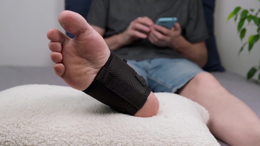 Man after an ankle injury is lying on the couch with his smartphone. close-up of the injured leg in special fixing bandage in the ankle joint area. rehabilitation after injury. close-up. slow motion. | Shutterstock HD Video #1089914371