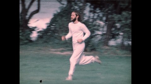 1970s: man runs through park in 1977 sportswear and fashion. Man with 1970s beard and long hair. Boy walks in park. Friends stand in park. Boy shouts to friend. Children run across park.