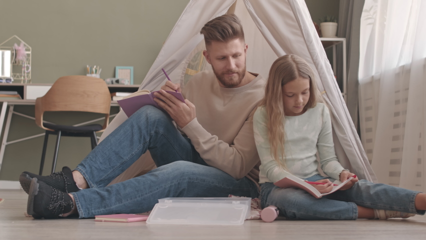 Slowmo of young Caucasian man with beard and his pretty 8 year old girl drawing in copybooks while sitting on floor in living room next to each other