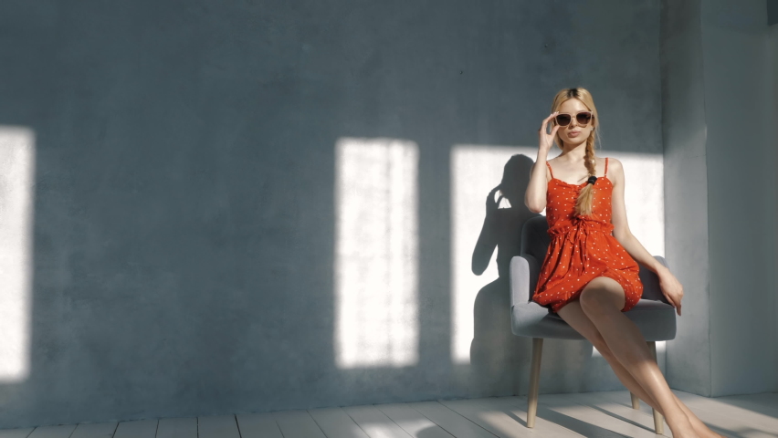 Young beautiful smiling female in trendy summer red dress. Sexy carefree blond woman sitting near wall in studio in a chair. Cheerful and happy in sunglasses. At sunny day. Shadow from window