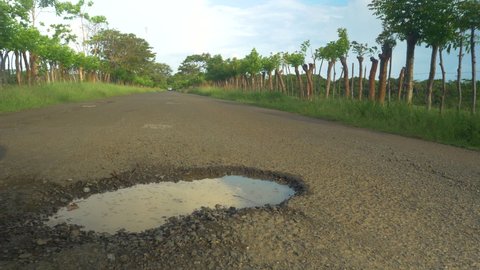CLOSE UP: Road pit filled with rainwater in the middle of a paved countryside road. Dangerous pothole with puddle in the middle of the asphalt road. Damage and bad road maintenance in third world.