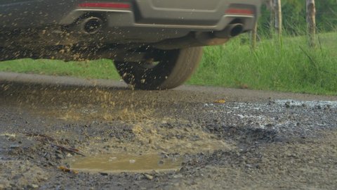 CLOSE UP: Car driving and splashing water from road pit puddle on paved road. Dangerous pothole with puddle in the middle of the asphalt road. Damage and bad road conditions in third world.
