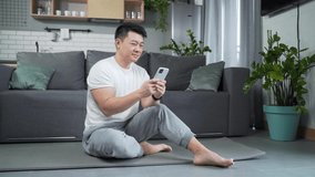 Athletic asian man sitting on yoga mat at home and using smartphone. Watch video tutorials. On the practice of yoga, training, workout. Fitness male browses mobile phone. indoor Happy athlete reads