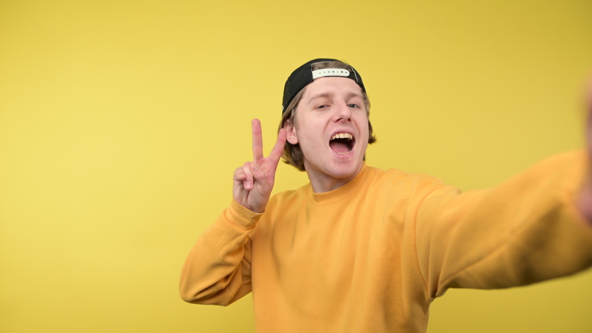 A happy guy in a cap and a yellow sweatshirt takes a selfie and shows a gesture of peace with a smile on his face, looking at the camera. Royalty-Free Stock Footage #1089924667