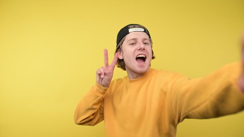 A happy guy in a cap and a yellow sweatshirt takes a selfie and shows a gesture of peace with a smile on his face, looking at the camera.