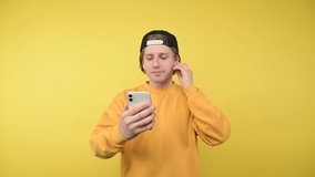 Positive guy in a yellow sweatshirt and cap makes a video call on a smartphone on a bright background, greets a friend and smiles.