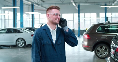 Handsome Caucasian young man in uniform and goggles talking on mobile phone in auto repair service. Male mechanic having conversation and chatting on telephone. Workday. Work concept.